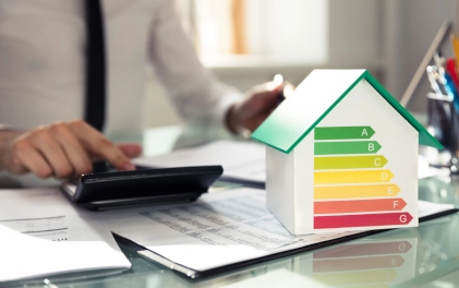 homme portant une chemise blanche et une cravate noire qui utilise une calculatrice sur un bureau en verre avec une maquette d'une maison avec le DPE