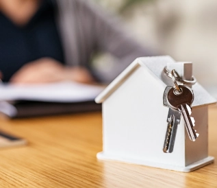 maquette d'une maison blanche avec les clés accrochées à la cheminée posée sur une table en bois avec une dame en fond