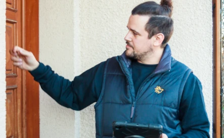 homme portant une doudoune sans manche Tuco qui frappe à la porte d'une maison et qui tient une tablette noire